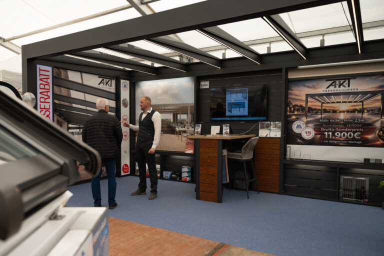 Two men talking inside an AKT Überdachungen trade show booth with a promotional poster and a screen nearby.