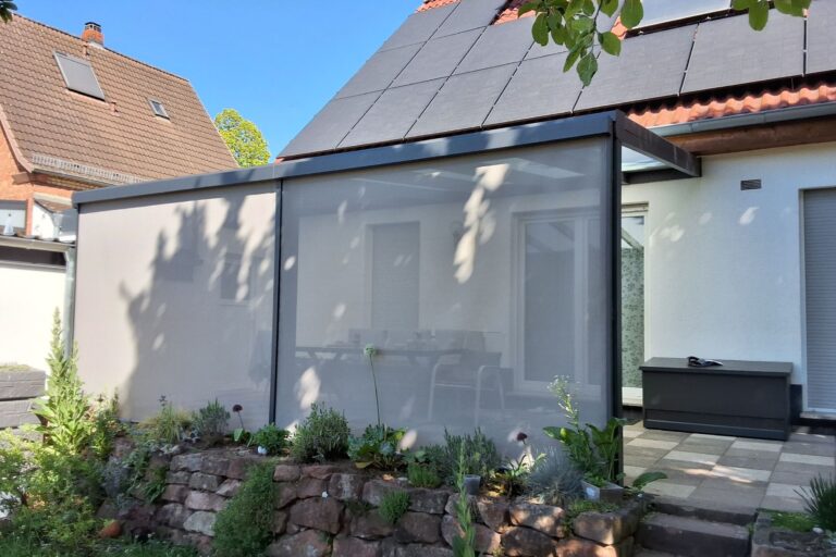 Modern outdoor patio with a frosted glass privacy screen and dark frame beside a garden bed and stone wall beneath a blue sky.