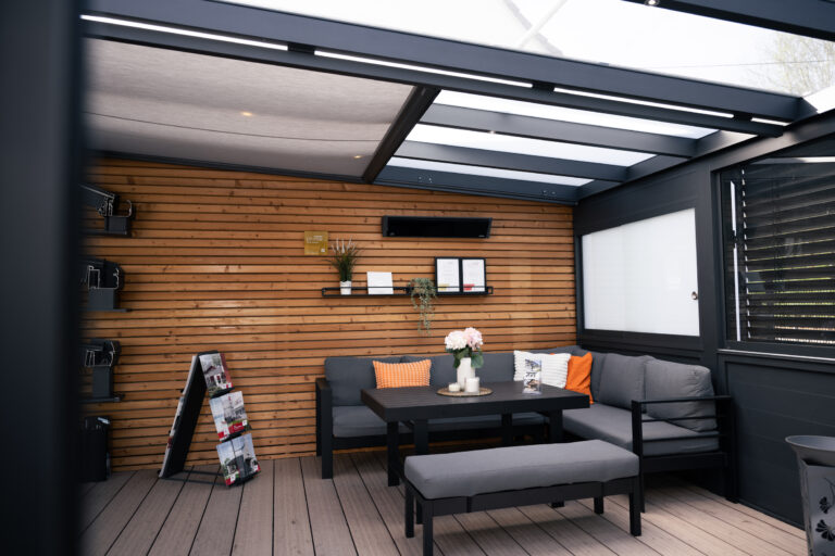 Outdoor lounge area with a wooden slat wall, gray sectional sofa, orange cushions, and a glass-roofed structure.