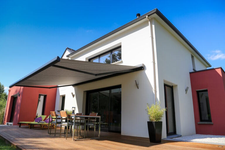 Modern white house with red accent wall, large glass doors, and a wooden deck with an outdoor dining set under a shaded pergola, blue sky above.