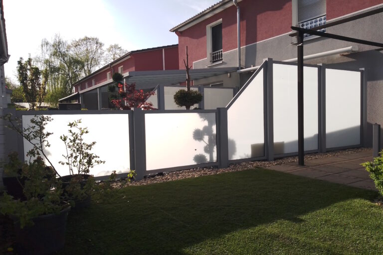 Modern courtyard with frosted glass privacy panels and a red-gray building, potted plants, gravel path and green lawn behind.