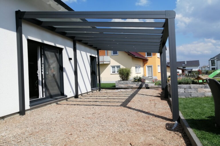 Modern house exterior with a metal pergola over a gravel patio and sliding glass doors; neighboring colorful houses in the background.