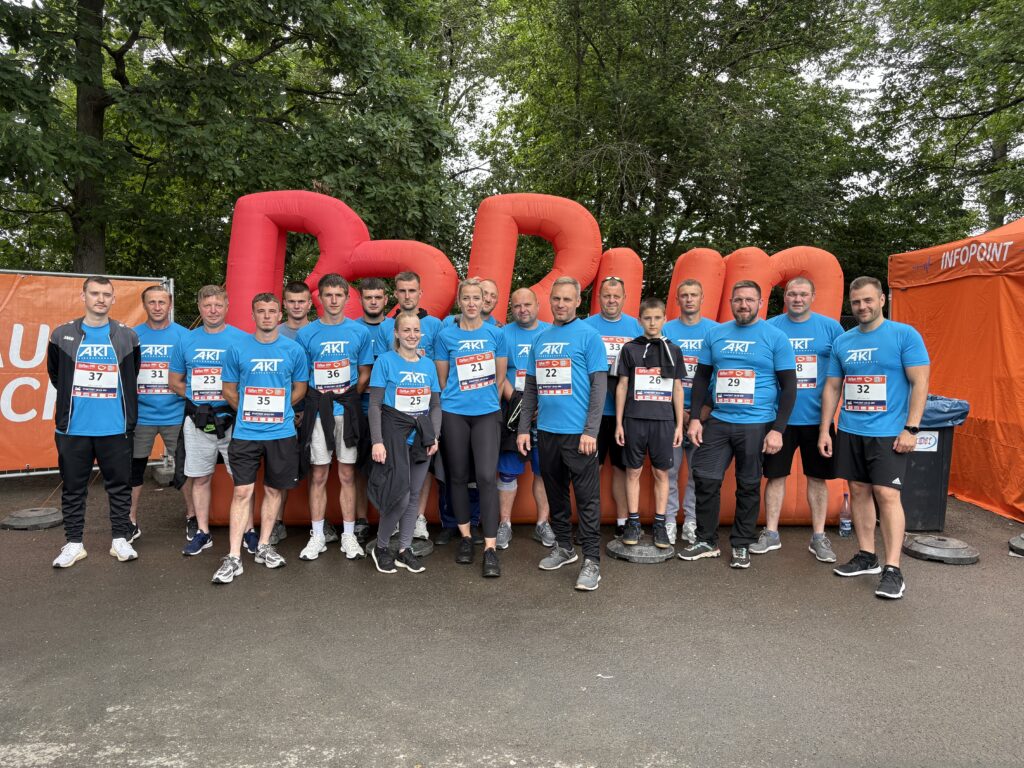 Group of runners in blue shirts with numbered bibs posing together for a photo at an outdoor race venue with an orange inflatable backdrop.
