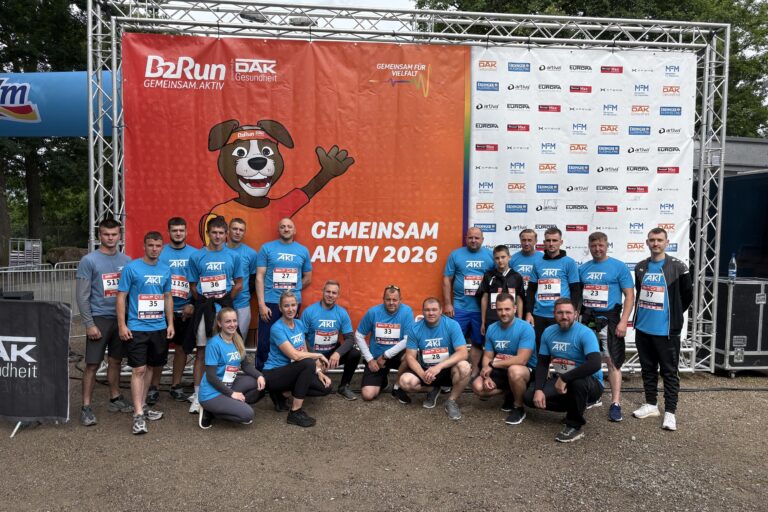 Group of people in blue t-shirts posing in front of a large orange event banner with a cartoon dog mascot and 'GEMEINSAM AKTIV 2026' text at an outdoor event site.